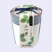 A white ceramic cup with blue dot patterning around the rim and a paper label that reads "Mitsuba Japanese Honeywort" with illustrations of leaves. A blue string bundles the label and the cup.