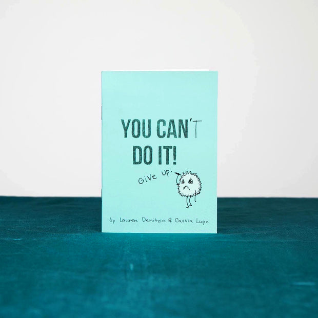 Small blue zine, "You Can't Do It!" with the "t" in can't being penciled in by a small fluffy cartoon creature, who also writes "Give up" under the title.