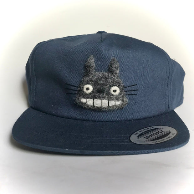 Navy blue flat bill snapback hat with a wool embroidery of Totoro's head, with a large smile on his face.