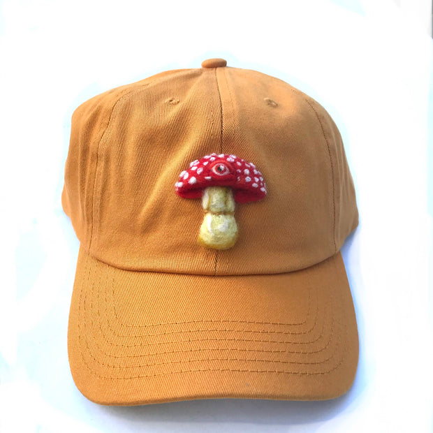 Yellow ochre colored curved bill cap with a 3D felted embroidery of a red mushroom with white spots and a single eyeball.