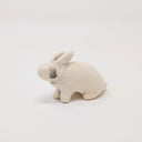 White ceramic rabbit, with simplistic body shapes and no facial features. It is positioned on all 4 paws, slightly sitting back with a gray smudged tear running from where its eye would be. Its ears are slightly back.