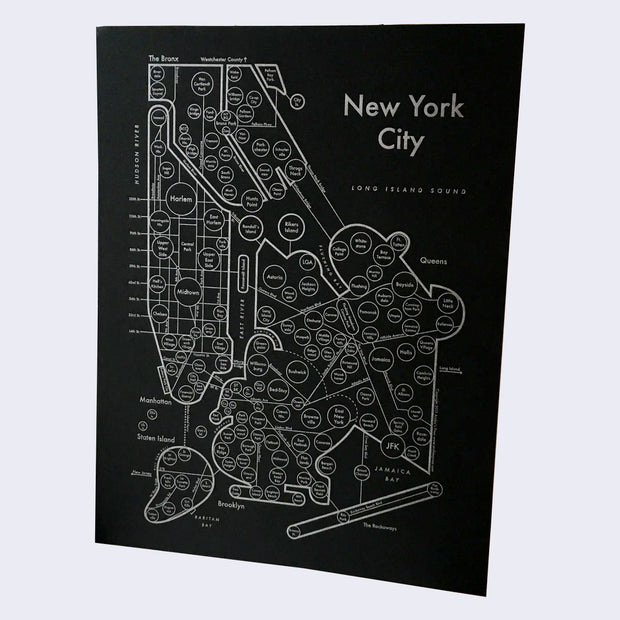 Silver letterpress on black paper of New York City, depicted abstractly as various circles, lines and abstract shapes. Neighborhood names are inside of circles, aligned in relation to their real location, and connected by street names. "New York City" is written largely in the upper right corner.