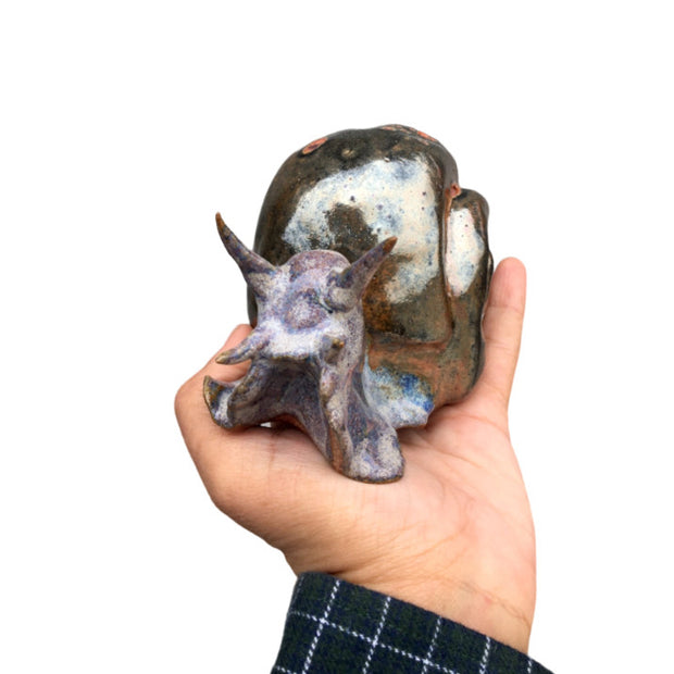 A ceramic snail being held in someone's hand, it has realistic features and is a purple color, with a bronze colored shell.