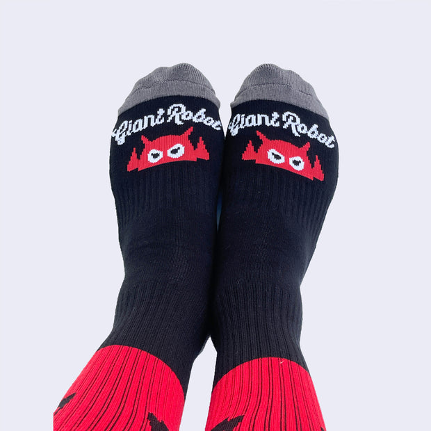 View of looking down at a model's feet wearing socks. The toe area has cursive text that say giant robot. Heel and toe area of sock is gray.