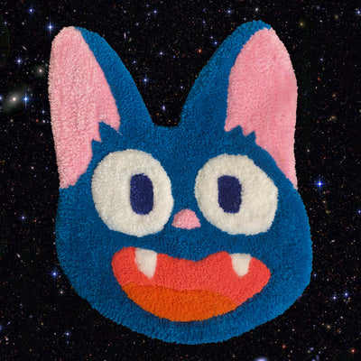 Tufted rug sculpture, of a blue smiling cat face akin to Jiji from Kiki's Delivery Service, mouth open smiling to show two pointy teeth and a red tongue.