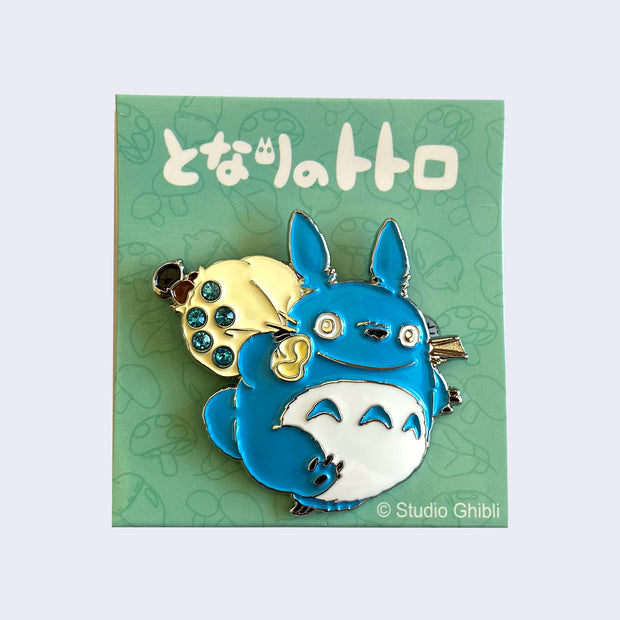 Brooch of a happy, chibi blue Totoro carrying a cream colored sack over its shoulder. There are 5 blue gems on the sack.