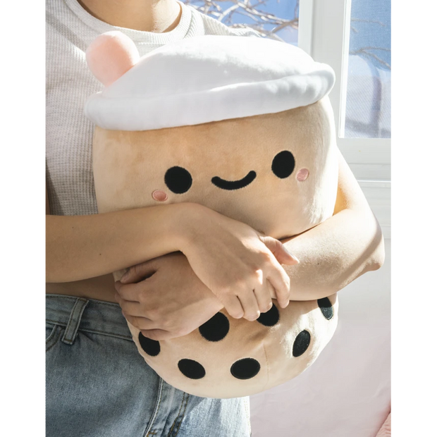 Rounded squishy brown plush shaped like a boba tea cup, with a cute kawaii smile and a pink straw coming out of the top. Being held in someones arms.