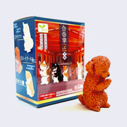 Small reddish brown plastic dog, fluffy and standing on its back paws. It has its head bowed and its hands folded together in prayer. It stands in front of its product packaging.