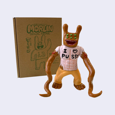 Brown painted soft vinyl figure, with 6 green eyes, a wide open mouth smile and two bunny like ears. The figure is muscular and wears a light pink shirt that says "I heart pussy" with two long tentacles as arms.