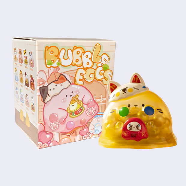 Yellow bubble shaped figure, semi transparent with yellow and white beads inside. It has a yellow cheeked white cat hat on its head and holds a daruma. It is in front of its packaging box, cream and pink with "Bubble Eggs" written. The side features an illustration of all design options.