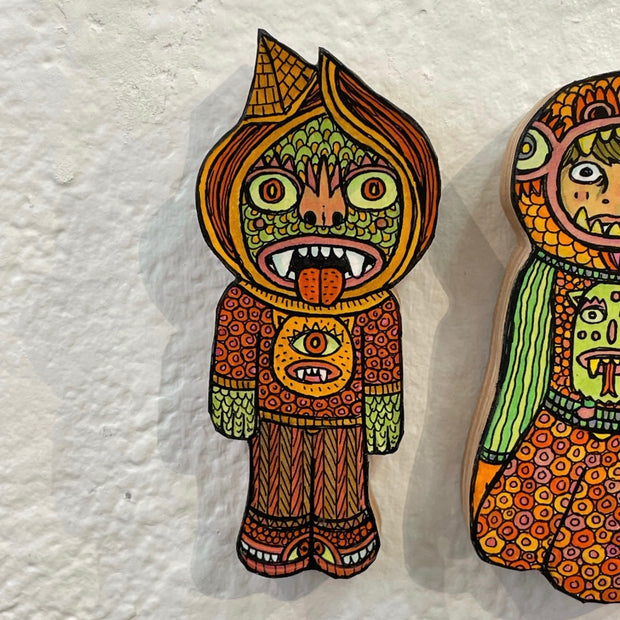 Small illustrated wood cut green goblin figure with its tongue out, an all orange outfit and a small pyramid as a hat. An illustration of a monster is on its sweater.
