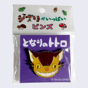 Enamel pin of a close up smiling Catbus face. On a purple backing card with Japanese writing, in a decorated plastic wrapping.