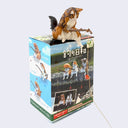 Small plastic fluffy Siberian cat, sitting on a box flat on its butt with its legs hanging down, holding a fishing pole made out of a thin branch. The box it sits on is its blind box packaging, which has photos of each different cat variation (tabby, calico, siberian cat, white with black and orange cat with folded ears).