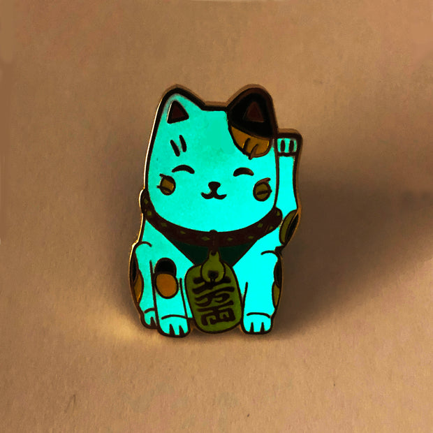 Enamel pin of a happy maneki cat, with yellow and black spots. Pin is glowing in the dark.