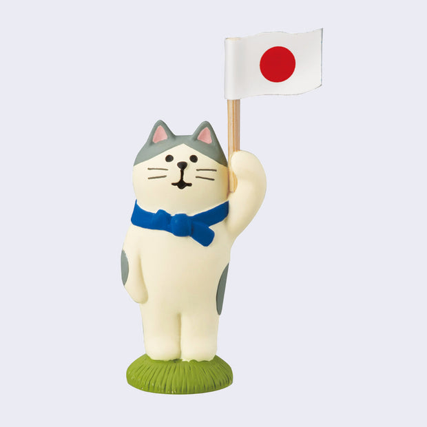 Small sculpture of white cat with gray spots, holding up a wooden post with a Japanese sticker flag on it. Cat wears a blue scarf and stands on a small green grass surface.