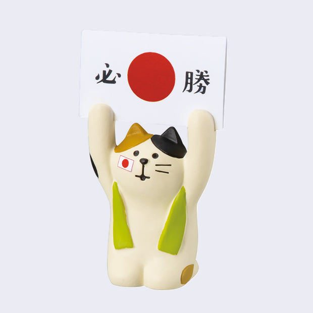 Small sculpture of a calico cat with a green scarf around its shoulders sitting on its knees and holding up a large card with the Japanese flag on it in both its paws.