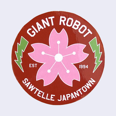 Burgundy circle sticker with "Giant Robot" written on top in white font and "Sawtelle Japantown" written on bottom. A pink cherry blossom is in middle with two green lightning bolds on each side.