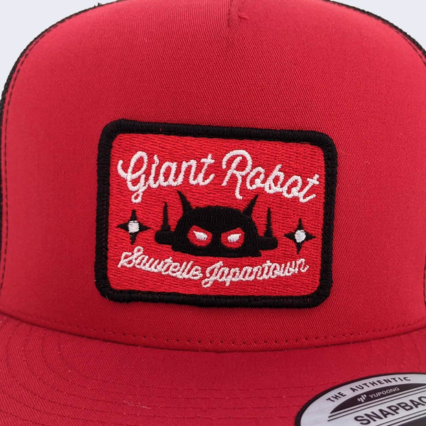 Close up of rectangle patch on the front of baseball cap. Entire patch is embroidered. Small black ninja stars decorate both sides of robot head.