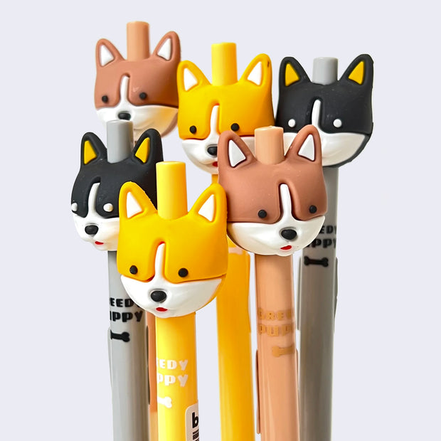 Arrangement of several retractable gel pens with rubber corgi heads as pen toppers. Pen body says "Greedy Puppy" with an illustration of a bone, with body color matching the puppy head. Colorways include: brown, yellow and black all with white snouts.