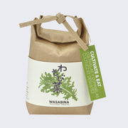  Brown paper sack, folded and tied with a paper tie and a green hang tag that reads "Cultivate & Eat". Wrapped around is a cream sheet, with an illustration of mustard greens and Japanese script in the middle. "Wasabina Mustard Greens" is written on the bottom.