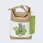 Brown paper sack, folded and tied with a paper tie and a green hang tag that reads "Cultivate & Eat". Wrapped around is a cream sheet, with an illustration of basil leaves and Japanese script in the middle. "Shiso Japanese Basil" is written on the bottom. 