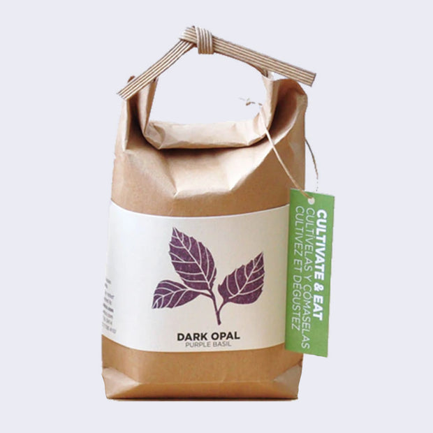 Brown paper sack, folded and tied with a paper tie and a green hang tag that reads "Cultivate & Eat". Wrapped around is a cream sheet, with an illustration of purple basil leaves. "Dark Opal Purple Basil" is written on the bottom.