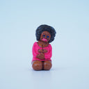 Small sculpture of a tan woman on her knees, with her hands in her lap. She wears a tight pink dress and has a perm.