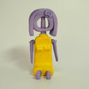 Sculpture of a purple character with no face (a hole is there instead) who sits on the ground in a yellow dress.