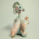 Clay sculpture of a woman with a deformed, sagging face and very large, stretched out boobs. She sits nude on the ground.