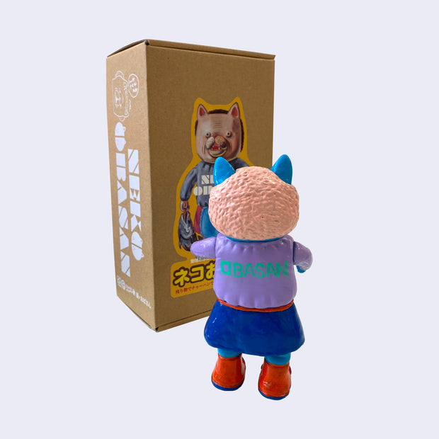 Back view of blue painted soft vinyl figure of a cat headed humanoid with light pink hair, wearing a purple sweater that says "obasan" on the back and a blue skirt with a red frilly apron. She holds a blue knife.