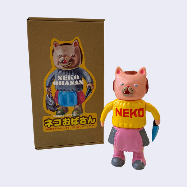 Light pink painted soft vinyl figure of a cat headed humanoid with brownish red hair, wearing a yellow sweater that says "neko" and a gray skirt with a pink frilly apron. She holds a blue knife.