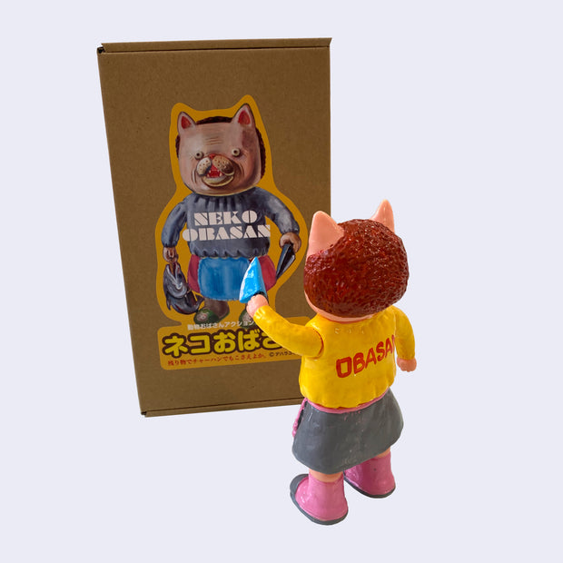Back view of light pink painted soft vinyl figure of a cat headed humanoid with brownish red hair, wearing a yellow sweater that says "Obasan" on the back and a gray skirt with a pink frilly apron. She holds a blue knife.
