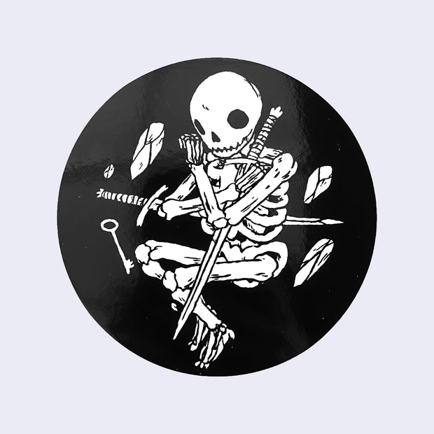 Black circle sticker with a drawing of skeleton clutching two swords. Small rocks and a key levitate around it.