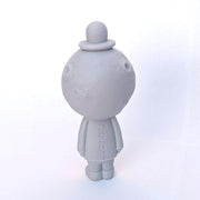 Lavender gray sculpture of a cratered moon with a simplistic, closed eye smiling face. It wears a bowler hat and a suit, arms at its side. Sculpture is reminiscent of Magritte.