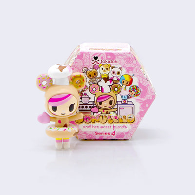 Small vinyl figure of a cartoon girl with pink bangs, wearing a hood with donuts for ears and a messy chef hat. Her arms are extended out, she has a sprinkle donut around her waist and a pink oven mitt. Behind is a hexagonal shaped pink Donutella and Her Sweet Friends (Series 4) display blind box.