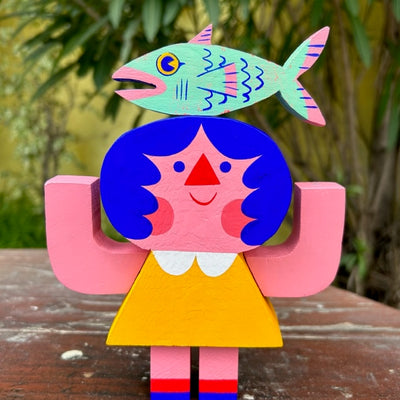 Die cut painted wooden sculpture of a pink girl with blue hair and simple features with her arms up at her side. She wears a yellow dress and balancing atop her head is a green and pink salmon fish.