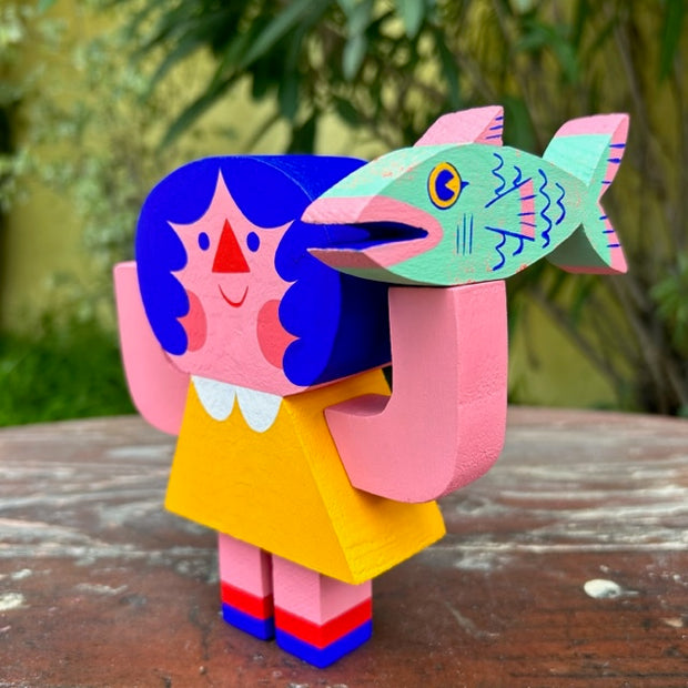Die cut painted wooden sculpture of a pink girl with blue hair and simple features with her arms up at her side. She wears a yellow dress and balances and green and pink salmon fish on her hand.