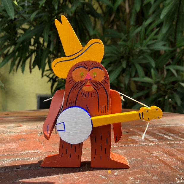 Die cut wood sculpture of a sweet looking brown fur Bigfoot, standing with a tall yellow cowboy hat and a banjo slung around its shoulders. Its arms are movable. 