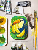 Painting of a blue toucan sitting atop of several bananas with a green grass.