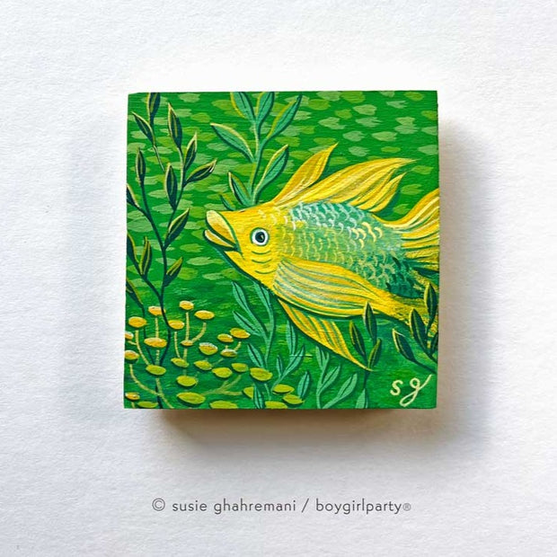 Painting on wooden panel of a yellow fish in a green underwater scene, with lots of simplified fish swimming behind and pieces of kelp nearby.