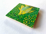 Painting on wooden panel of a yellow fish in a green underwater scene, with lots of simplified fish swimming behind and pieces of kelp nearby.