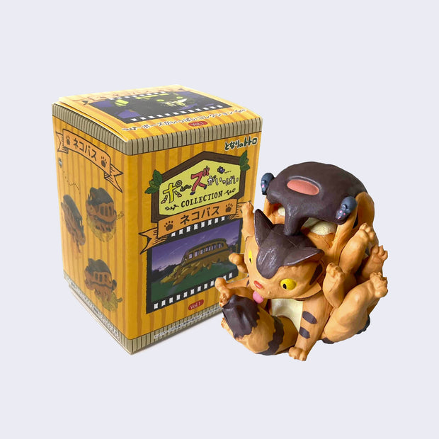 Vinyl figure of Catbus from My Neighbor Totoro, curled up over itself to lick its tail. It stands in front of a yellow striped product box, with Japanese script and a still from My Neighbor Totoro.
