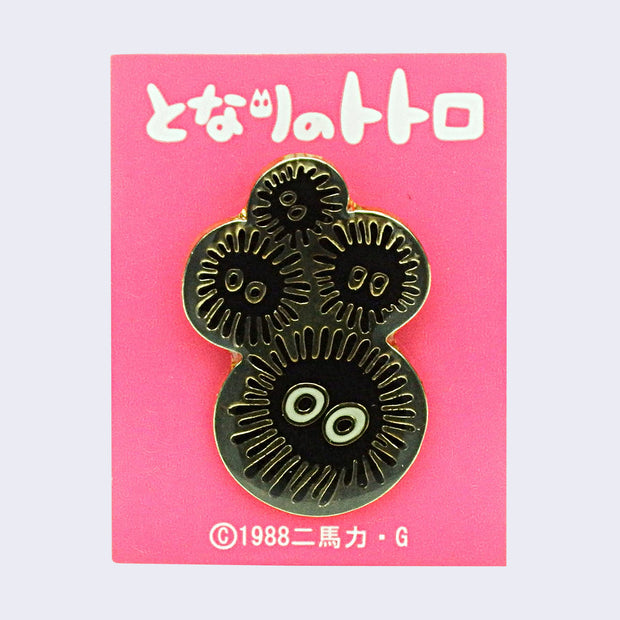 Enamel pin of 4 black circular Soot Sprites atop one another, with a gold border. On a pink backing card with Japanese writing.