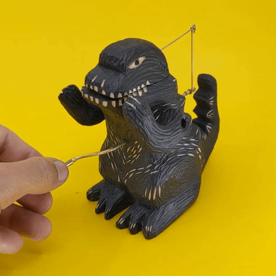 Carved wooden sculpture of a black Godzilla, standing with arms propped up and crank on its stomach. Gif of crank being turned, opening and closing its mouth.