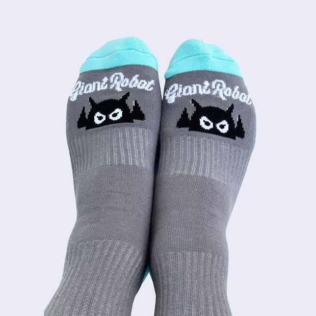 Front view of model's feet wearing gray socks. An angry cartoon robot head is on instep. White cursive text above robot says giant robot.
