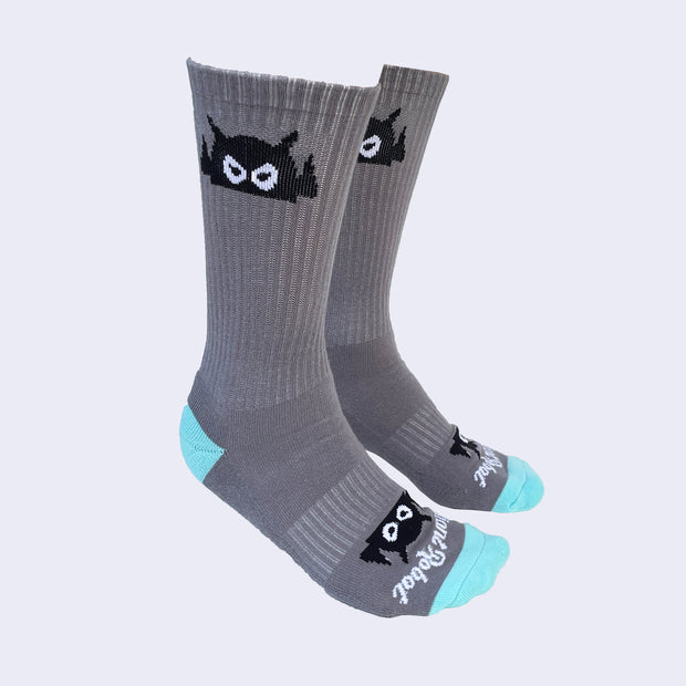 A side view of model's feet wearing gray socks decorated with a cartoon robot head on the cuffs. Toes and heel area is a bright turquoise color.