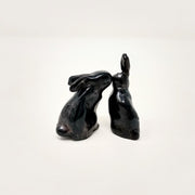 Two black ceramic rabbits with simplistic body shapes and no facial features, one sits on its hind legs with its ears straight up. The other has their ears put back and leans in, appearing to whisper in the other's ear.