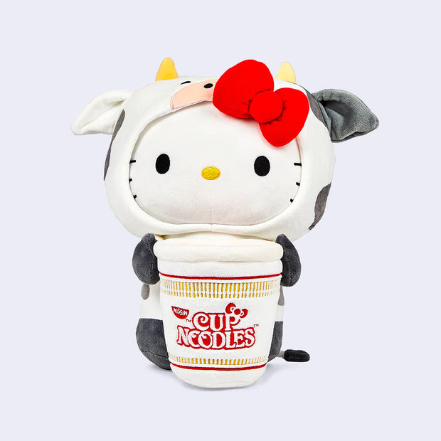 Plush version of Hello Kitty dressed in a cow suit with small horns and her red bow atop her head. She has her arms wrapped around a large Nissin Cup of Noodles.