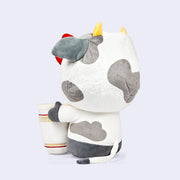 Plush version of Hello Kitty dressed in a cow suit with small horns and her red bow atop her head. She has her arms wrapped around a large Nissin Cup of Noodles. Back angle.
