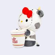 Plush version of Hello Kitty dressed in a cow suit with small horns and her red bow atop her head. She has her arms wrapped around a large Nissin Cup of Noodles. Side angle.